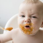 Mother feeding messy baby son with spoon in high chair